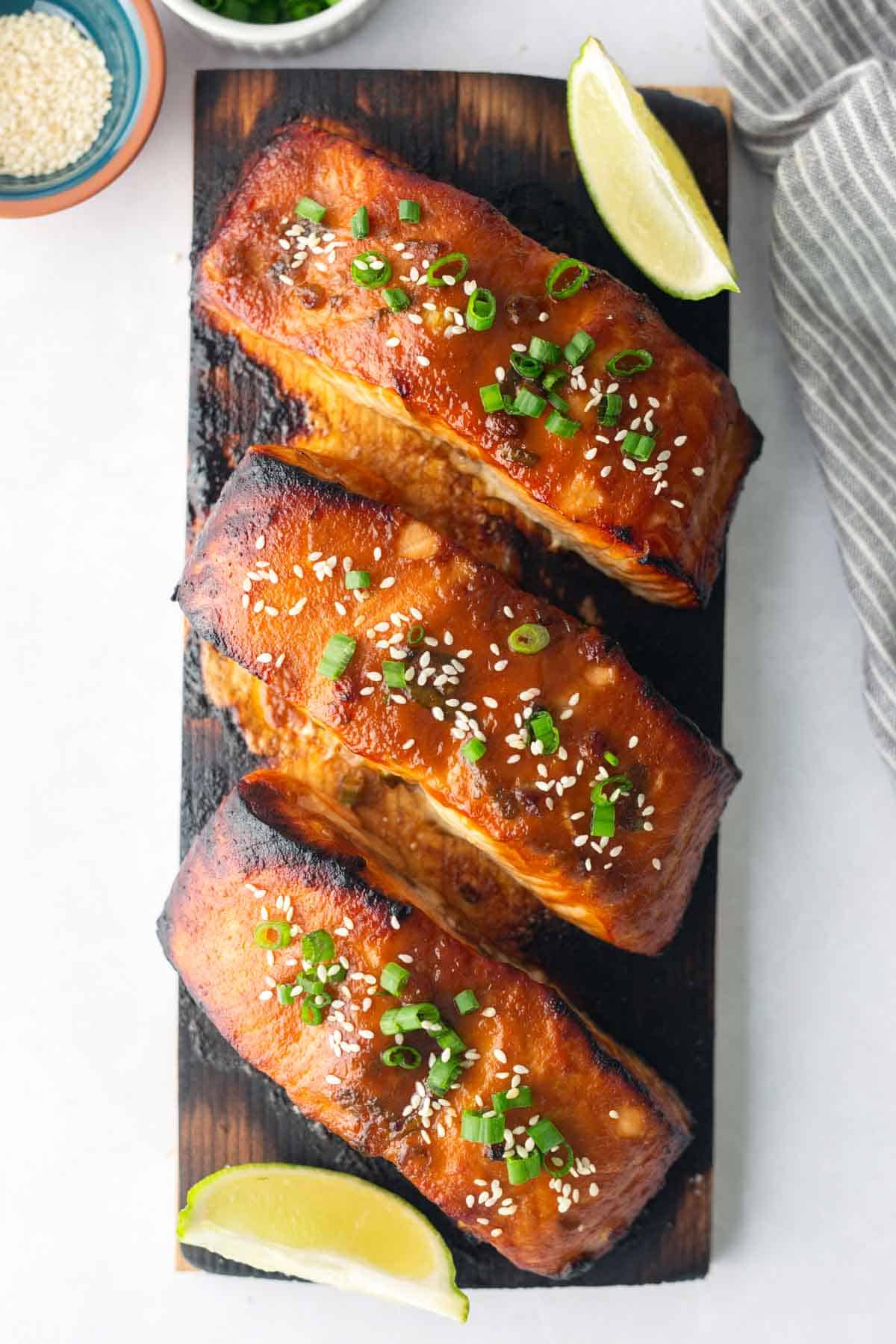 Three pieces of cedar plank salmon, glazed to perfection and garnished with sesame seeds and chopped green onions, are arranged on a wooden board with fresh lime wedges.