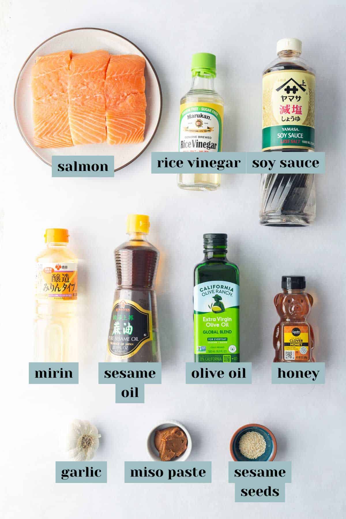 Top-down view of ingredients on a white surface labeled: cedar plank salmon, rice vinegar, soy sauce, mirin, sesame oil, olive oil, honey, garlic, miso paste, and sesame seeds.
