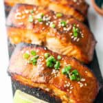 Three glazed cedar plank salmon fillets topped with sesame seeds and chopped green onions are arranged on a wooden board with a lime wedge in front.