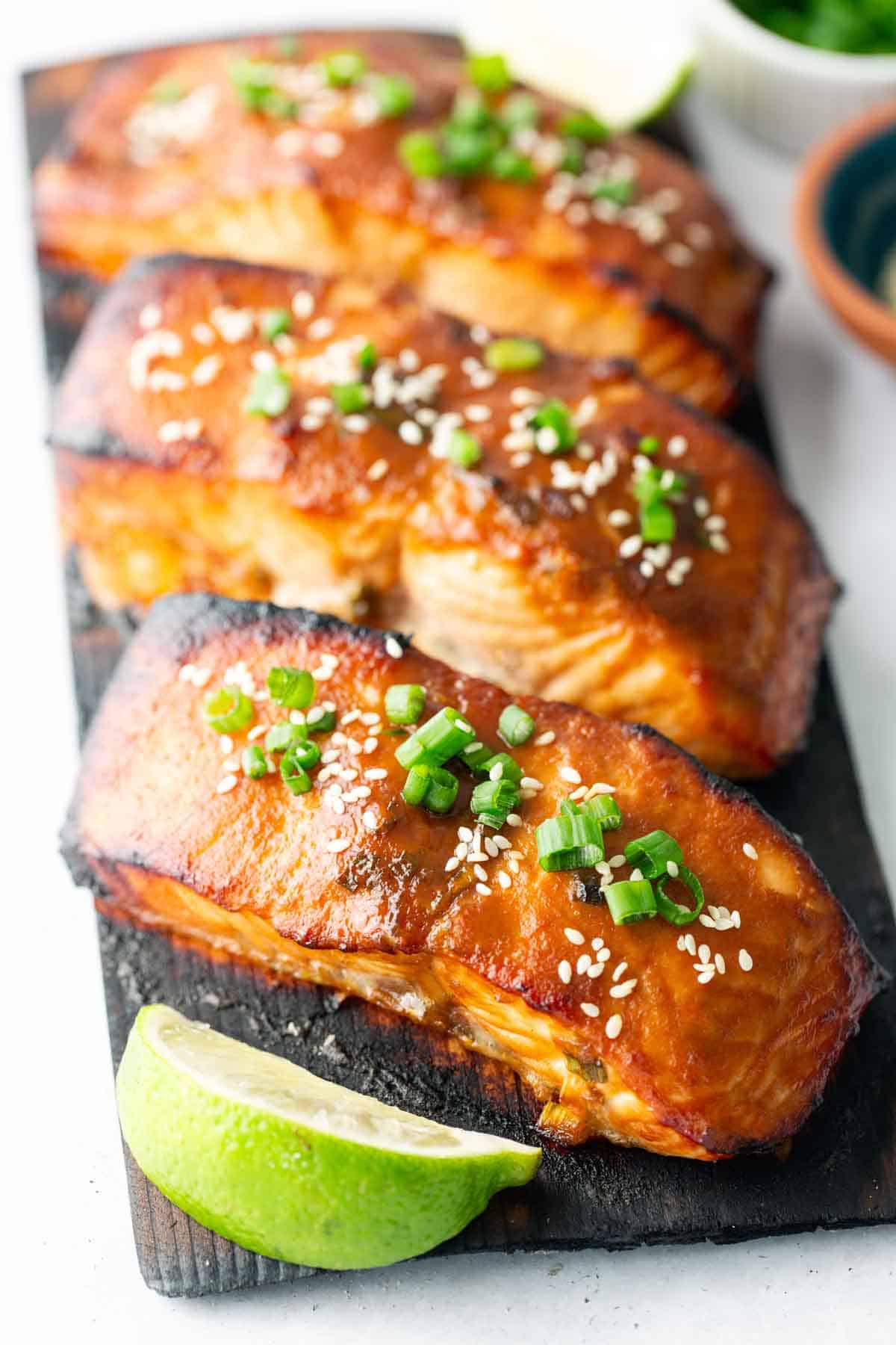 Three glazed cedar plank salmon fillets topped with sesame seeds and chopped green onions are arranged on a wooden board with a lime wedge in front.