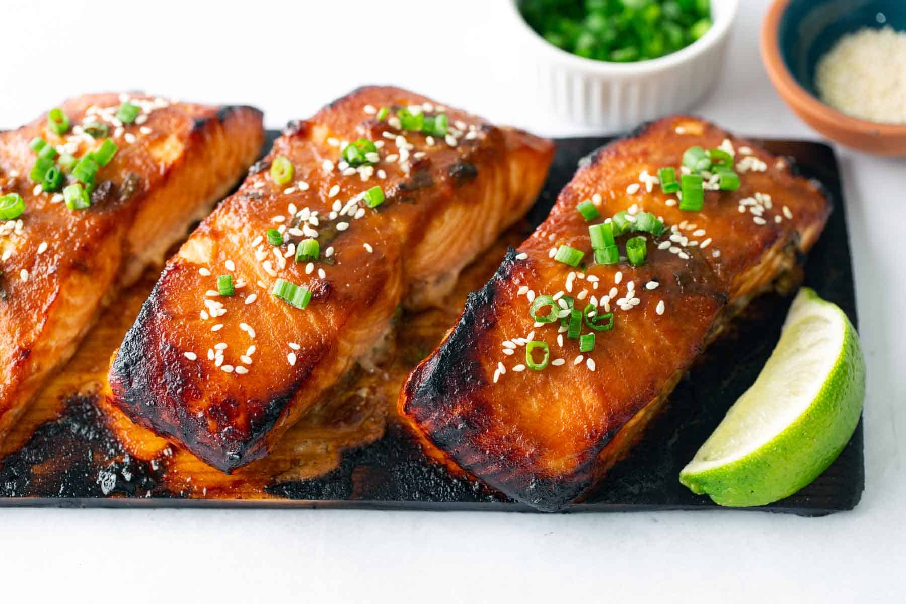 Three pieces of cedar plank salmon glazed to perfection, topped with sesame seeds and chopped green onions, served on a wooden board with a lime wedge. Small bowls of green onions and sesame seeds in the background.