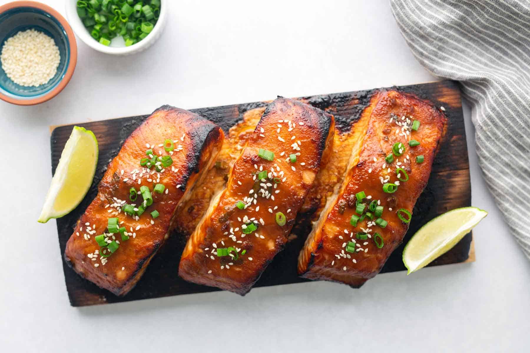 Three grilled cedar plank salmon fillets garnished with sesame seeds and chopped green onions, served on a wooden board with lime wedges. Small bowls of sesame seeds and green onions are nearby.