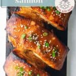 Three glazed salmon fillets on a cedar plank, garnished with sesame seeds and green onions, with lime wedges on the side.