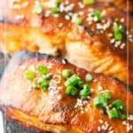 Close-up of cedar plank salmon fillets topped with sesame seeds and chopped green onions, served on a wooden plank with a lime wedge nearby.