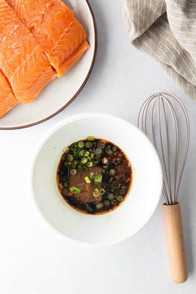 Raw salmon fillets on a plate, reminiscent of preparation for cedar plank salmon, sit beside a bowl of sauce with green onions and a whisk on a white surface.