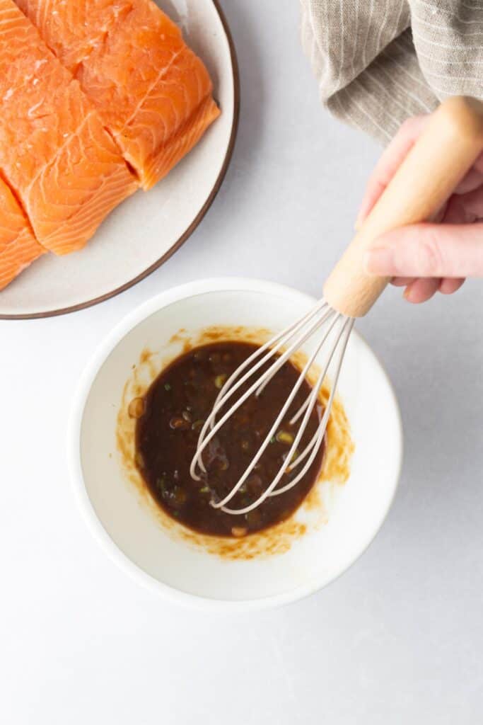 A hand whisks a dark marinade in a white bowl next to a plate with raw salmon fillets, preparing them for cedar plank salmon.