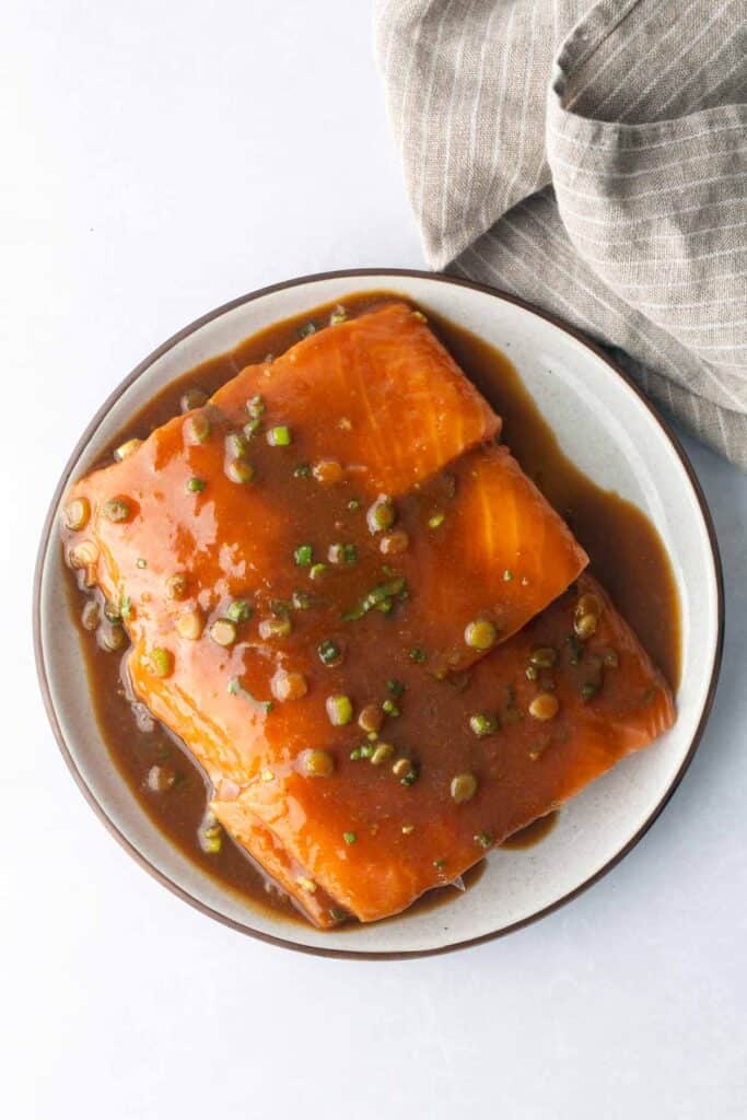 Two raw salmon fillets sit on a plate, ready to become cedar plank salmon, covered in a brown marinade with chopped green onions. A gray striped cloth is visible in the upper right corner.