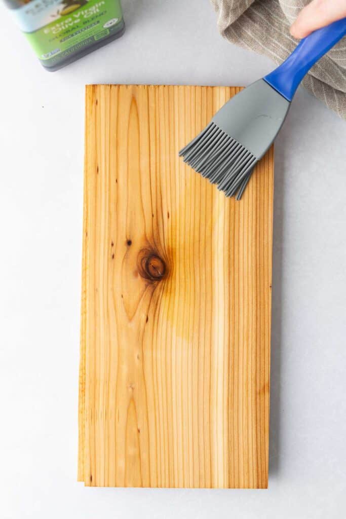 A cedar plank, intended for cedar plank salmon, is being brushed with oil using a silicone brush. A bottle of oil and a towel are partially visible in the background.
