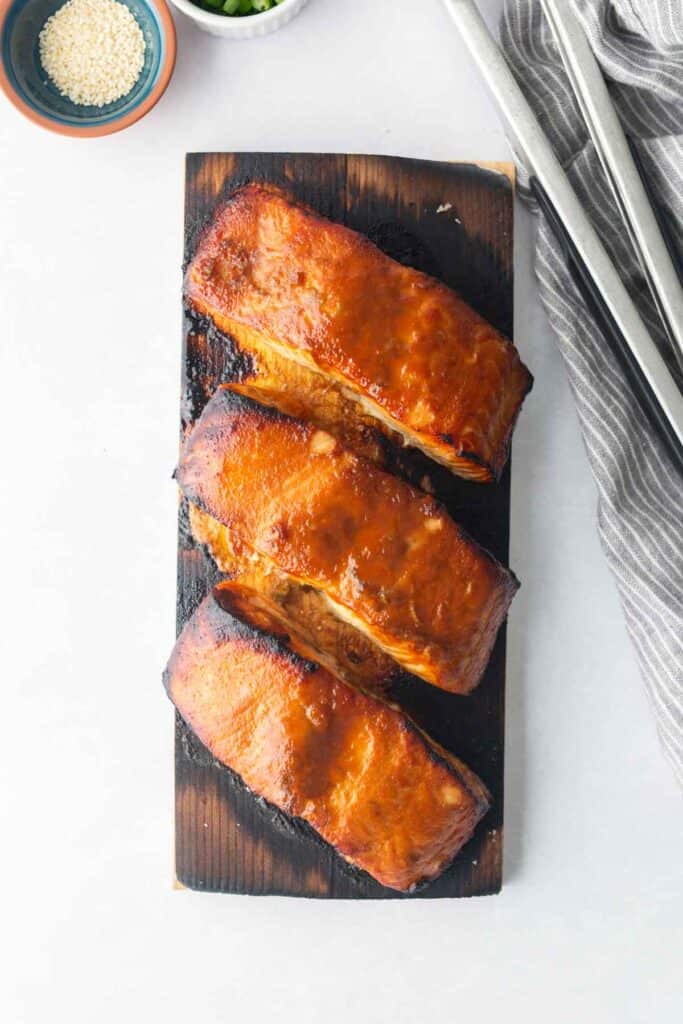 Three glazed cedar plank salmon fillets on a wooden plank, photographed from above, with a bowl of sesame seeds and striped cloth nearby.