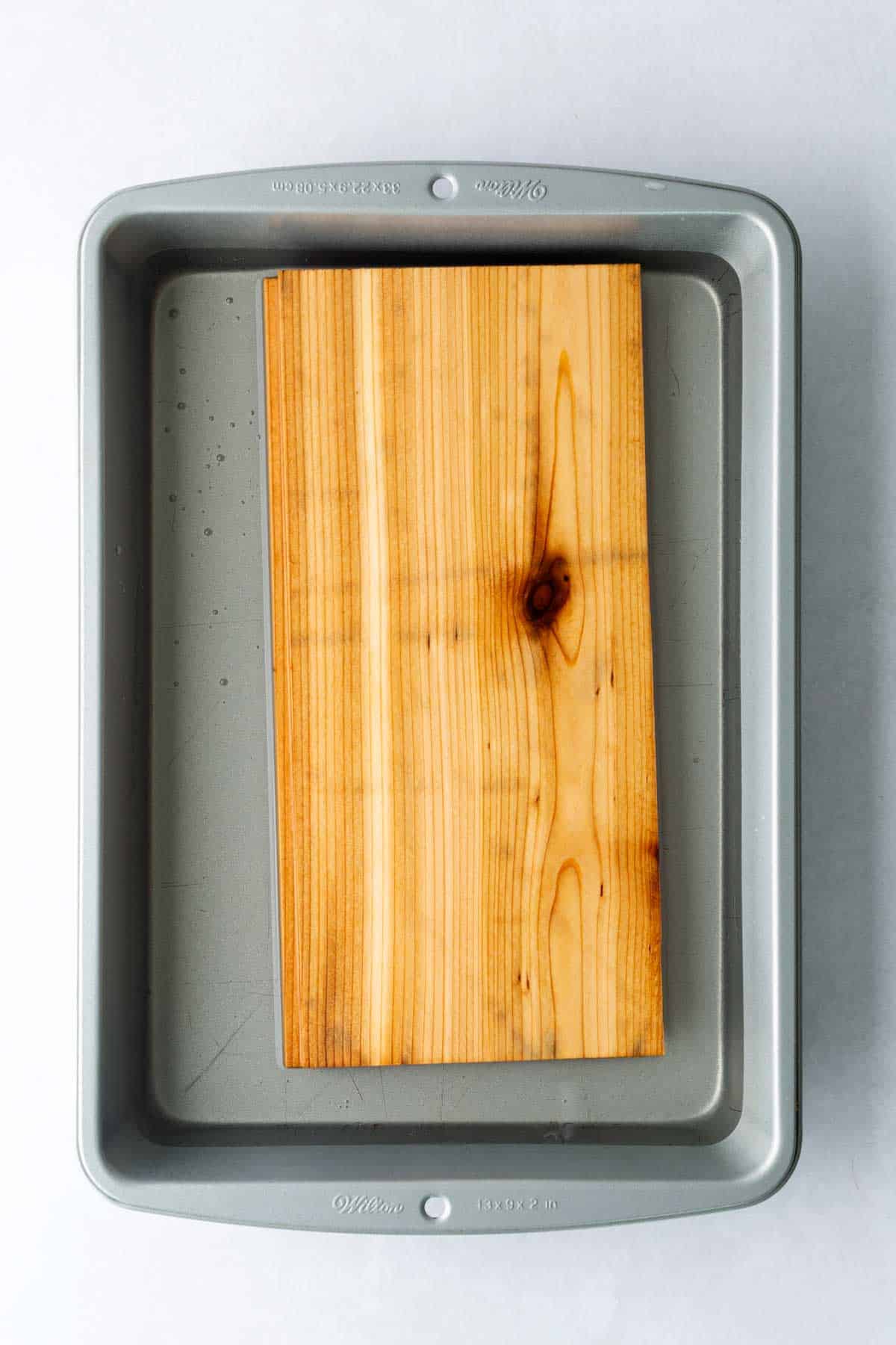 A rectangular cedar plank, ready for making cedar plank salmon, sits inside a metal baking pan on a light surface.