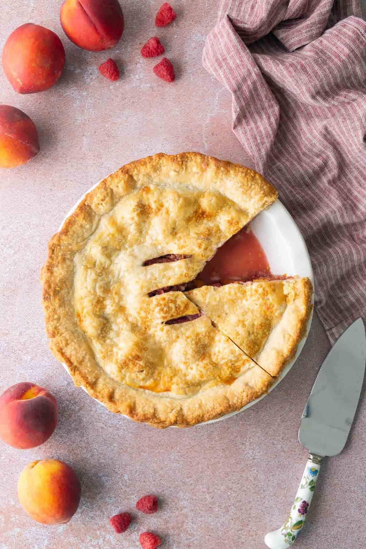 Peach Raspberry Pie will brighten up your summer days. Peach and raspberry pie with one slice missing, surrounded by whole peaches, raspberries, a pie server, and a pink cloth on a light surface.