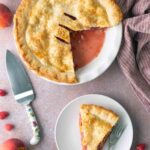 A peach and raspberry pie with one slice cut out, shown next to a plate with a single slice, surrounded by fresh peaches and raspberries on a textured surface.