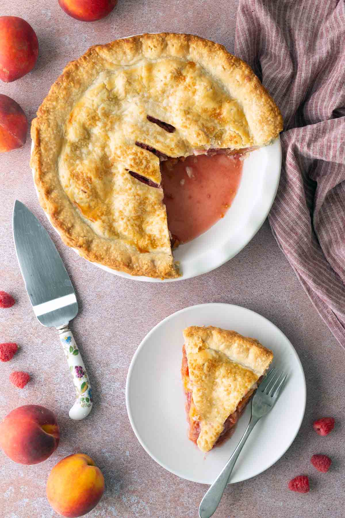 A peach and raspberry pie with one slice cut out, shown next to a plate with a single slice, surrounded by fresh peaches and raspberries on a textured surface.