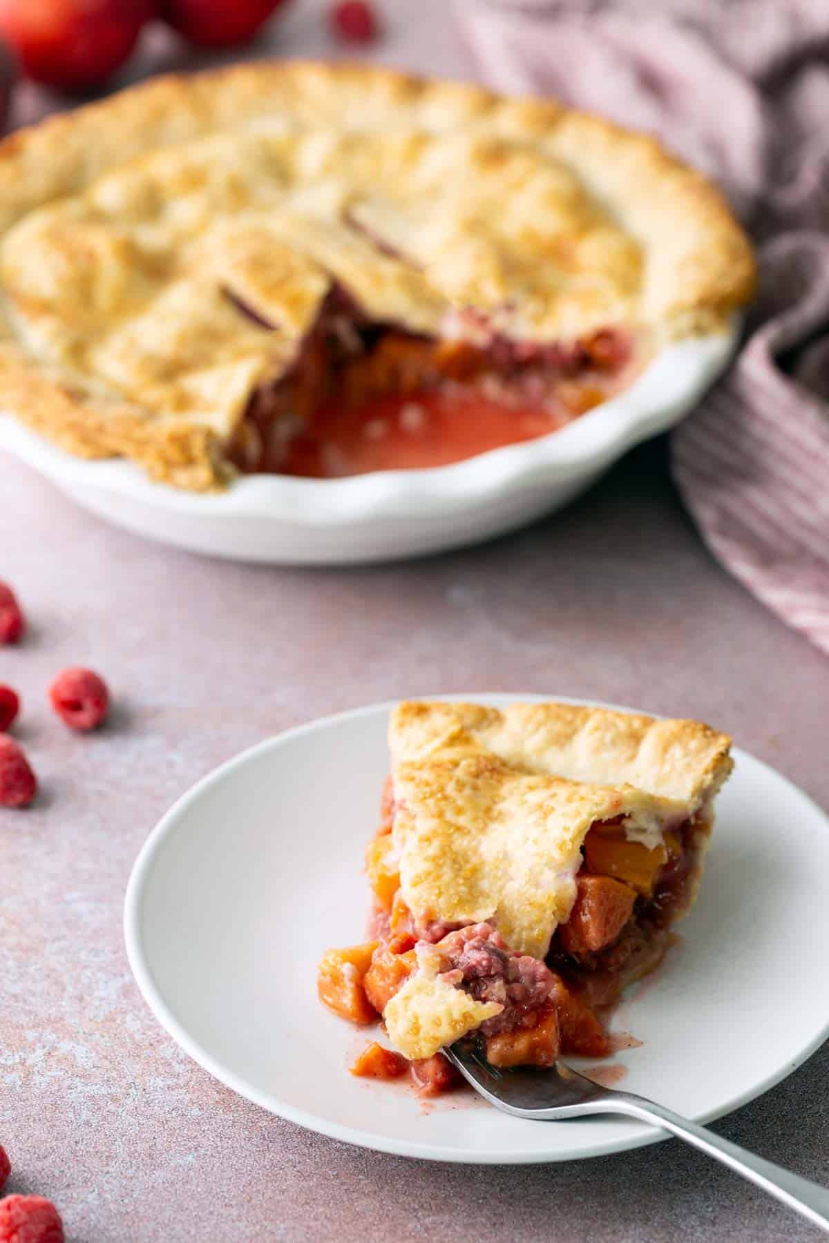 A slice of fruit pie with golden crust on a white plate, with whole pie and scattered raspberries in the background.
