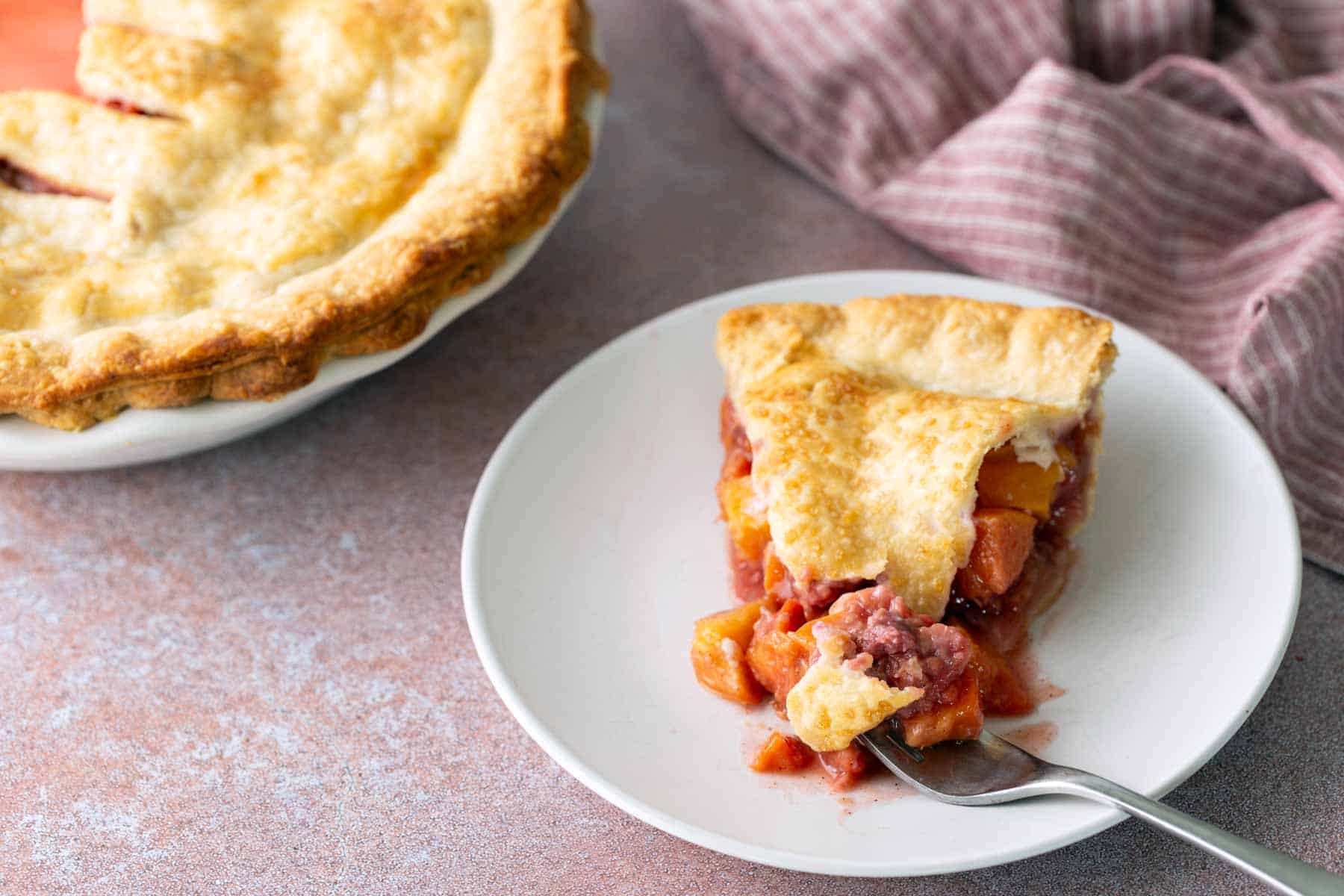 A slice of fruit pie with a golden crust on a white plate, with a fork and a larger pie and striped cloth in the background.