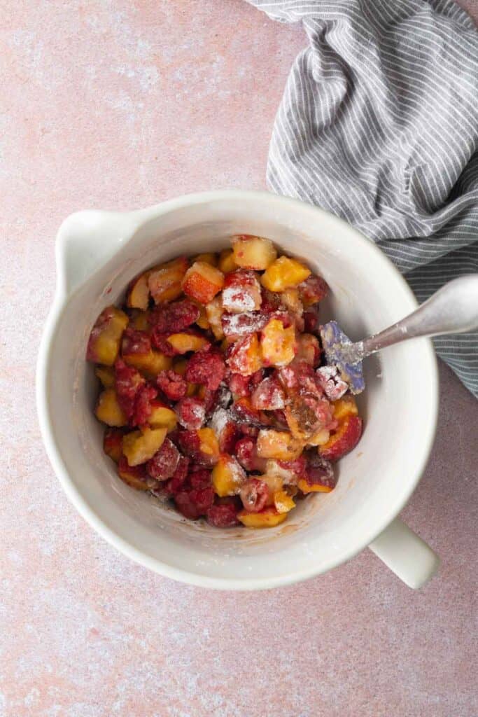 White mixing bowl with chopped peaches and raspberries being mixed with a spoon, next to a gray and white striped cloth on a pink surface.