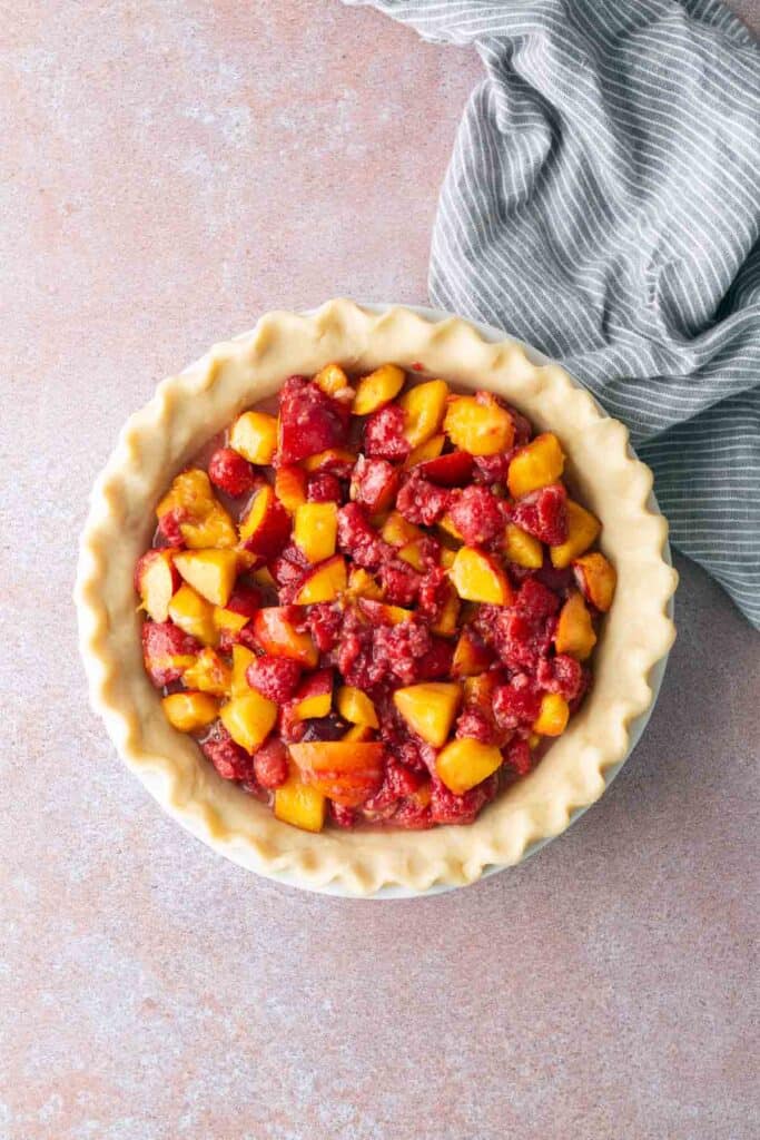 Unbaked pie with a lattice crust edge filled with a mixture of diced peaches and strawberries, placed on a light surface next to a gray and white striped cloth.