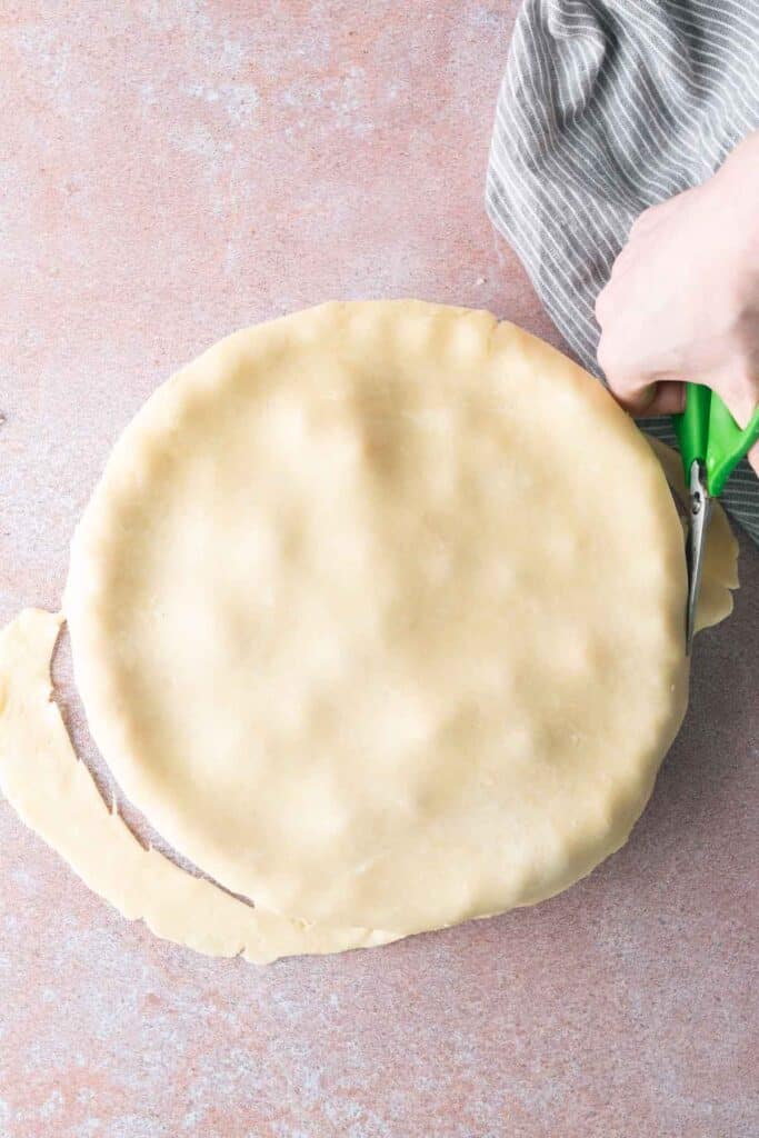 A hand uses scissors to trim excess pie dough from the edge of a round pie on a light brown surface.