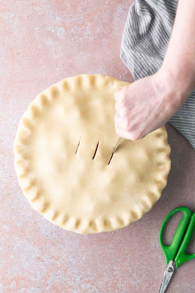 A hand is cutting three slits into the top crust of an unbaked pie next to a pair of green scissors on a light-colored surface.