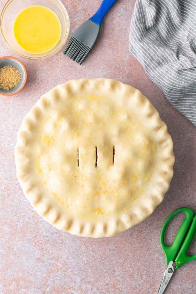 Top-down view of an unbaked pie with a fluted crust and three steam vents, surrounded by a pastry brush, egg wash, brown sugar, a cloth, and scissors on a pink surface.