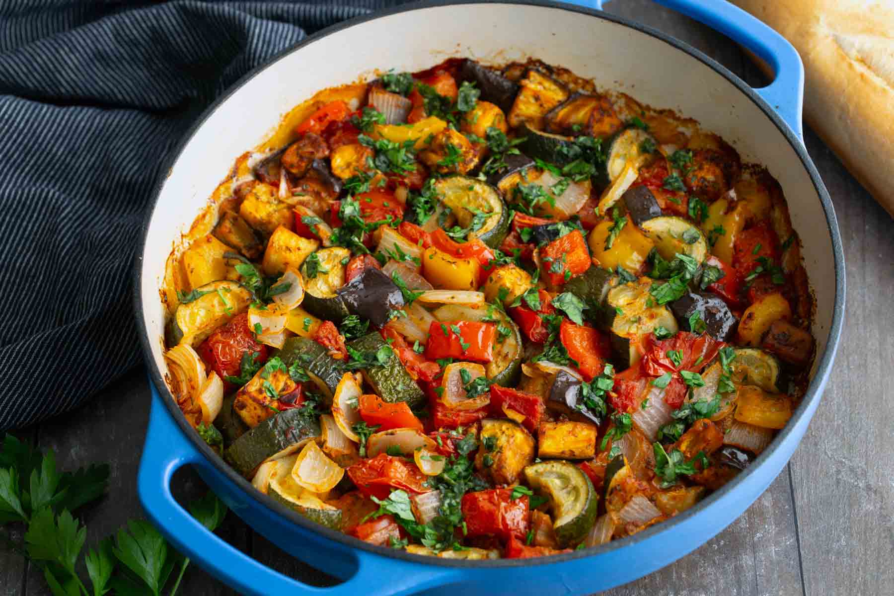 A blue pot filled with cooked ratatouille made of chopped vegetables like zucchini, tomatoes, eggplant, onion, and herbs sits on a wooden table next to a loaf of bread.