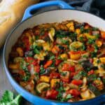 Blue pot filled with cooked ratatouille, featuring chopped vegetables like zucchini, tomatoes, and peppers, on a wooden table with a loaf of bread, parsley, and a dark cloth nearby.