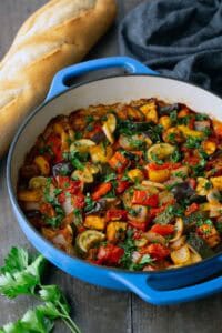 Blue pot filled with cooked ratatouille, featuring chopped vegetables like zucchini, tomatoes, and peppers, on a wooden table with a loaf of bread, parsley, and a dark cloth nearby.