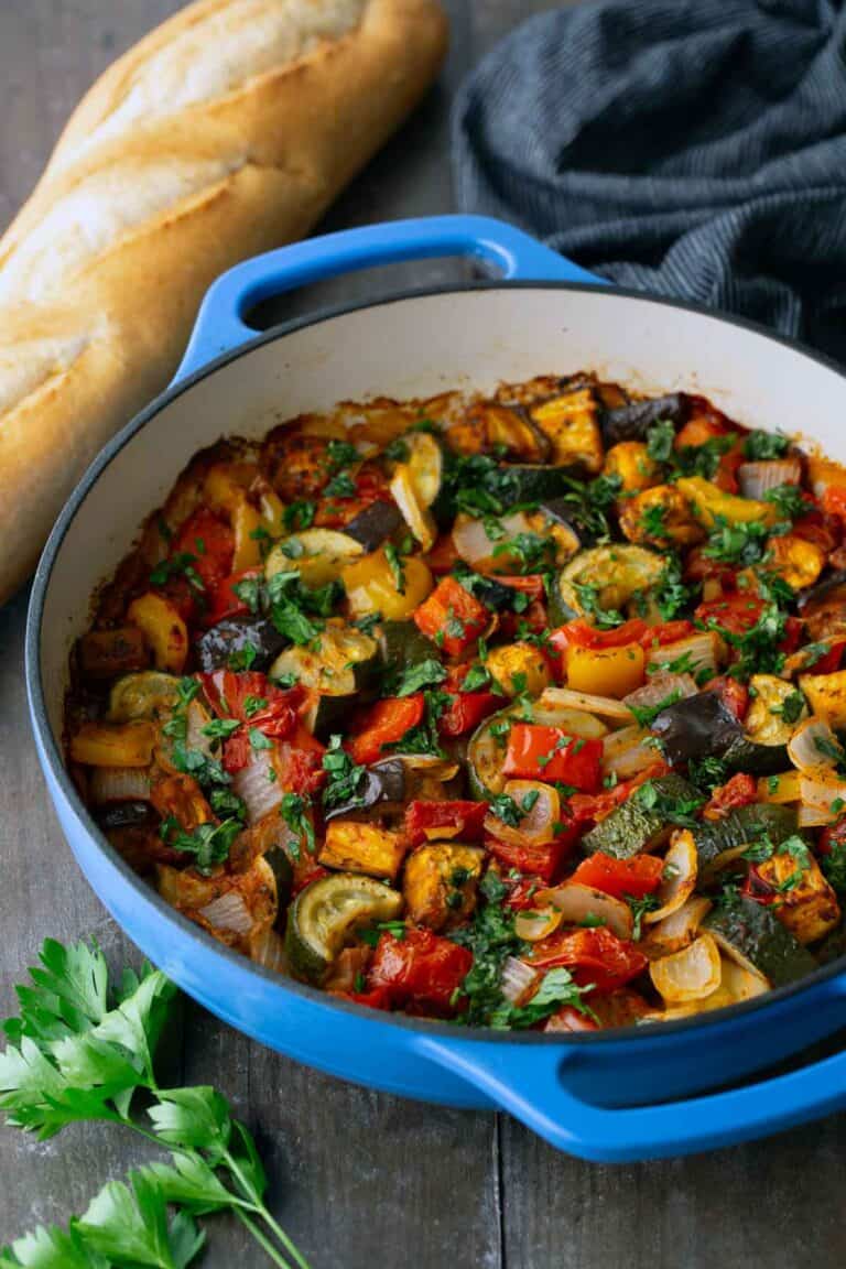 Blue pot filled with cooked ratatouille, featuring chopped vegetables like zucchini, tomatoes, and peppers, on a wooden table with a loaf of bread, parsley, and a dark cloth nearby.