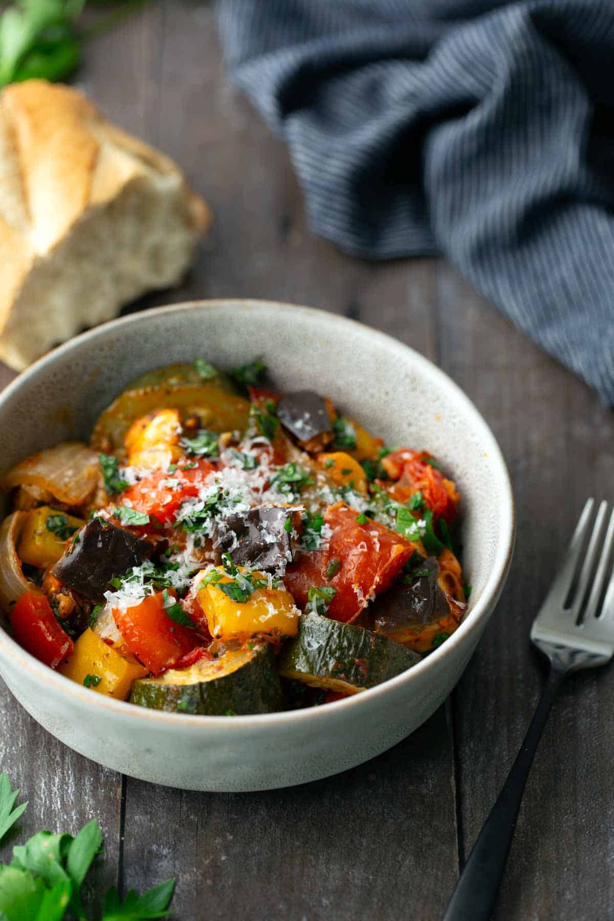 A bowl of ratatouille with chopped vegetables and grated cheese, placed on a wooden table next to a fork, bread, and a striped cloth.