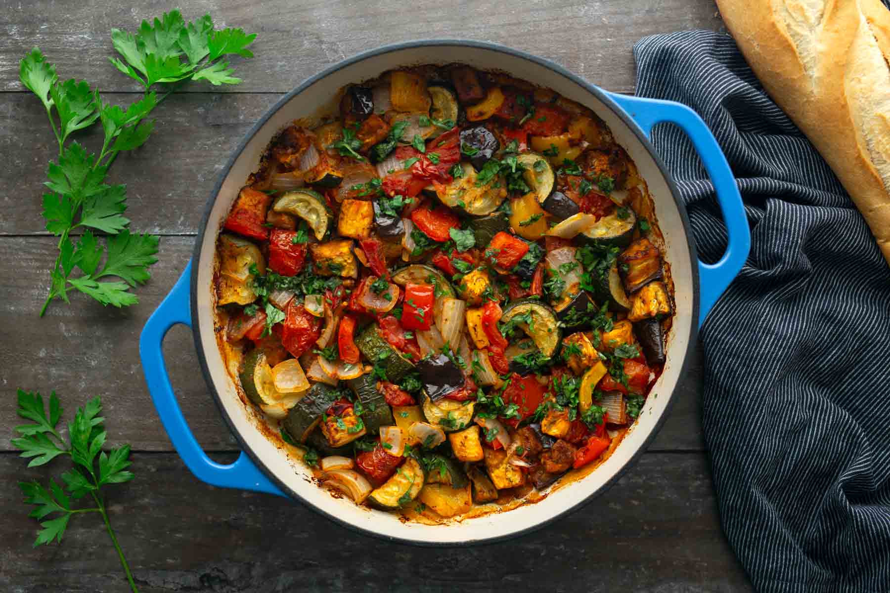 A blue pot filled with ratatouille made of chopped vegetables sits on a wooden table, next to parsley, a cloth napkin, and a loaf of bread.