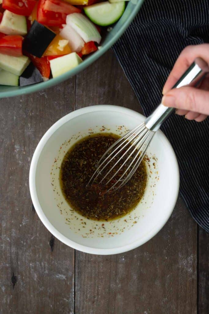 A hand whisks a bowl of herb vinaigrette on a wooden table, next to a bowl of chopped vegetables and a striped cloth.