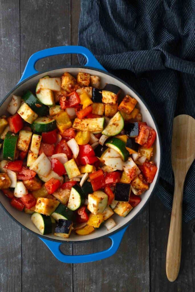 A blue pot filled with diced, seasoned vegetables including zucchini, bell peppers, onion, eggplant, and tomatoes sits on a wooden surface next to a wooden spoon and a striped cloth.