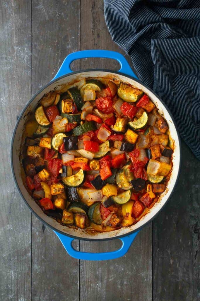 Blue pot filled with cooked ratatouille, including zucchini, eggplant, tomatoes, onions, and red peppers, on a dark wooden table next to a folded striped cloth.