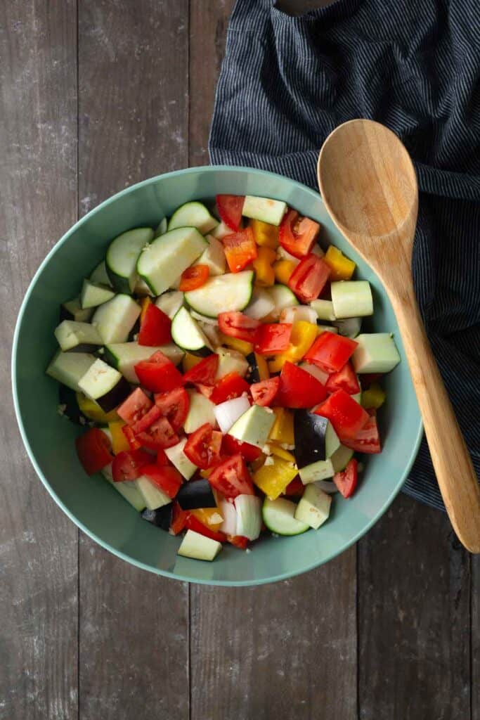 A green bowl filled with chopped tomatoes, cucumbers, bell peppers, and onions sits on a wooden surface next to a wooden spoon and a striped cloth.