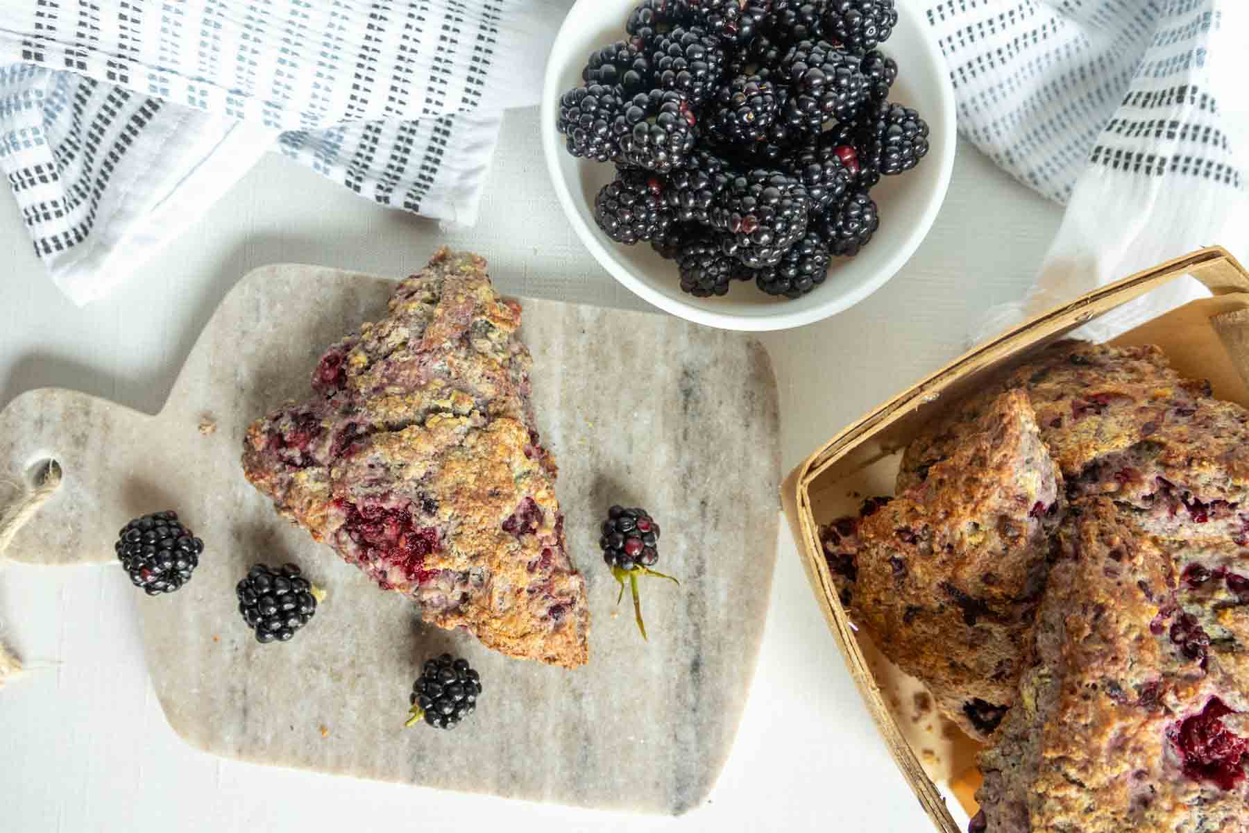 A blackberry scone on a marble board, fresh blackberries nearby, with more scones in a basket and blackberries in a white bowl.