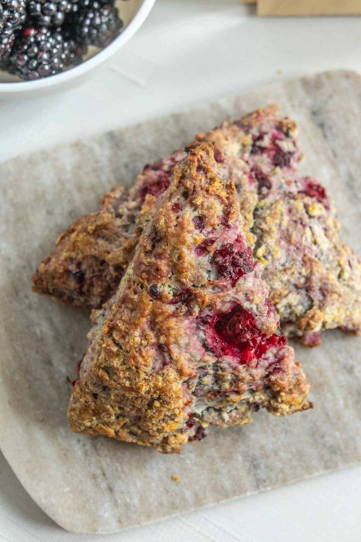 Two triangular blackberry scones with visible berry pieces rest on a light stone slab, with a bowl of blackberries in the background.