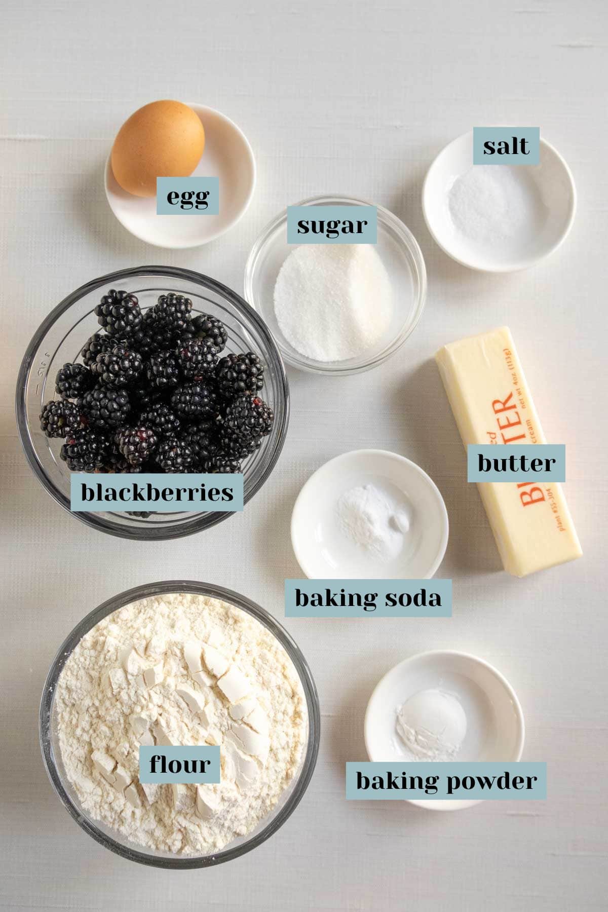 Ingredients on a white surface: an egg, sugar, salt, blackberries, butter, flour, baking soda, and baking powder, each labeled in small bowls.