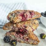 Three blackberry scones are stacked on a marble board, with fresh blackberries scattered nearby and a bowl of berries in the background.