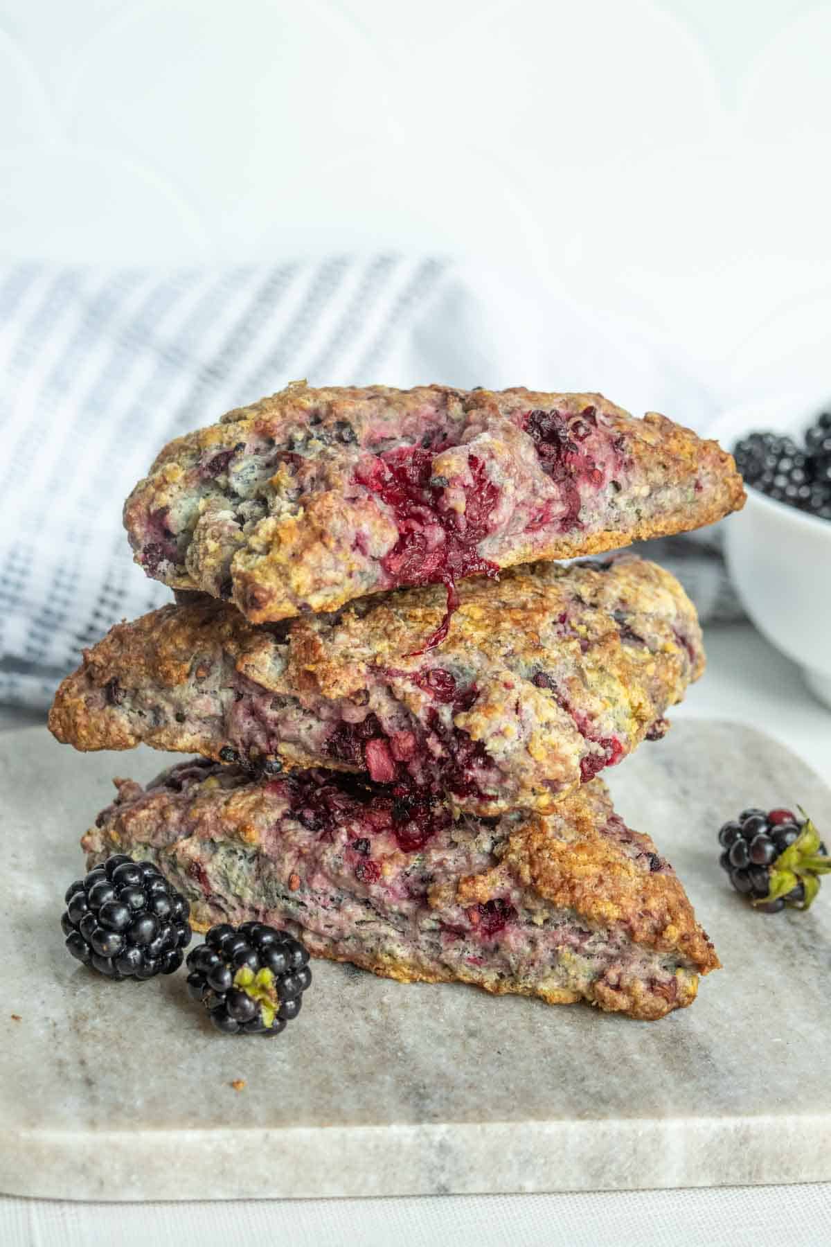 Three blackberry scones are stacked on a marble board, with fresh blackberries scattered nearby and a bowl of berries in the background.