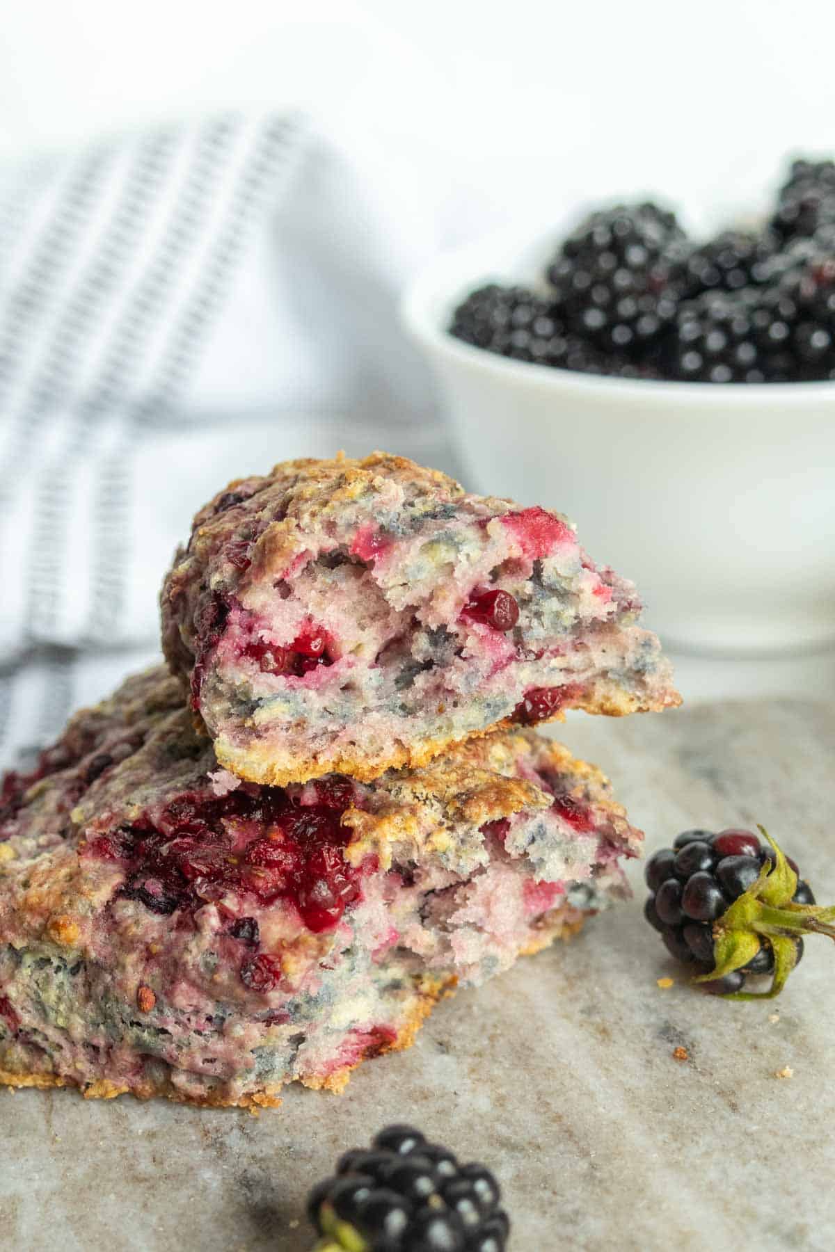 Two berry scones rest on a stone surface, one split open to reveal a colorful interior. A white bowl of blackberries and a striped towel sit in the background. Blackberries are scattered nearby.