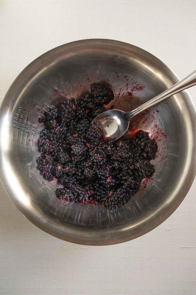 Blackberries and juice in a stainless steel mixing bowl with a metal spoon, placed on a light-colored surface.