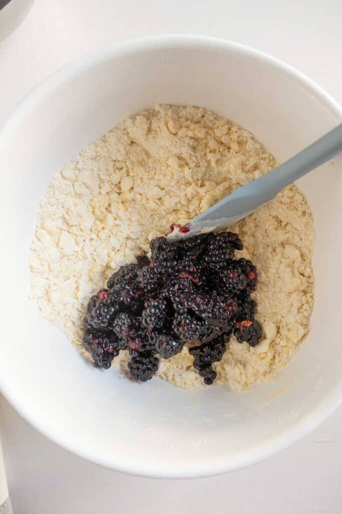 A white mixing bowl containing flour mixture and fresh blackberries, with a spatula partially visible.