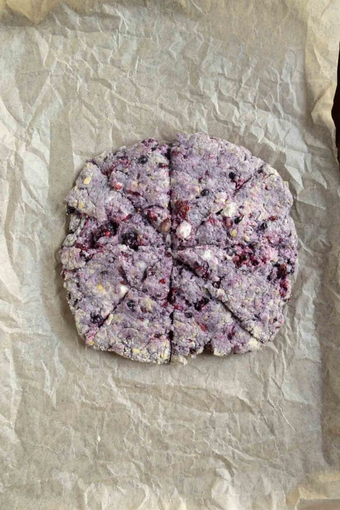 Unbaked, round blueberry scone dough cut into wedges sits on crumpled parchment paper, ready for baking.