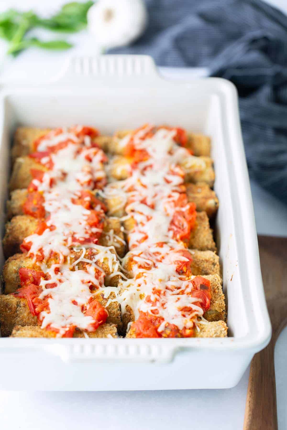 A white baking dish filled with breaded vegetable sticks topped with tomato sauce and melted cheese, placed on a white surface next to a wooden spatula.