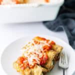 A white plate with two breaded, baked stuffed shells topped with tomato sauce and melted cheese, next to a fork. A casserole dish with more of the same food is in the background.