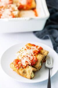 A white plate with two breaded, baked stuffed shells topped with tomato sauce and melted cheese, next to a fork. A casserole dish with more of the same food is in the background.