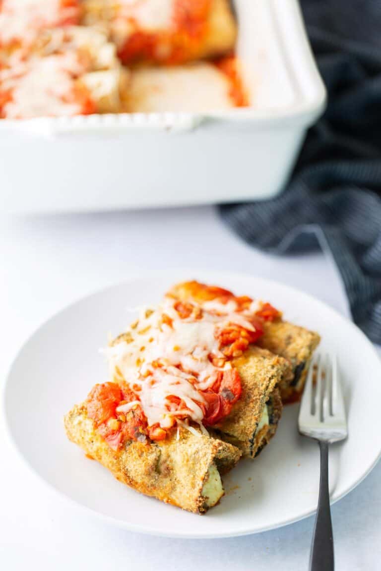 A white plate with two breaded, baked stuffed shells topped with tomato sauce and melted cheese, next to a fork. A casserole dish with more of the same food is in the background.