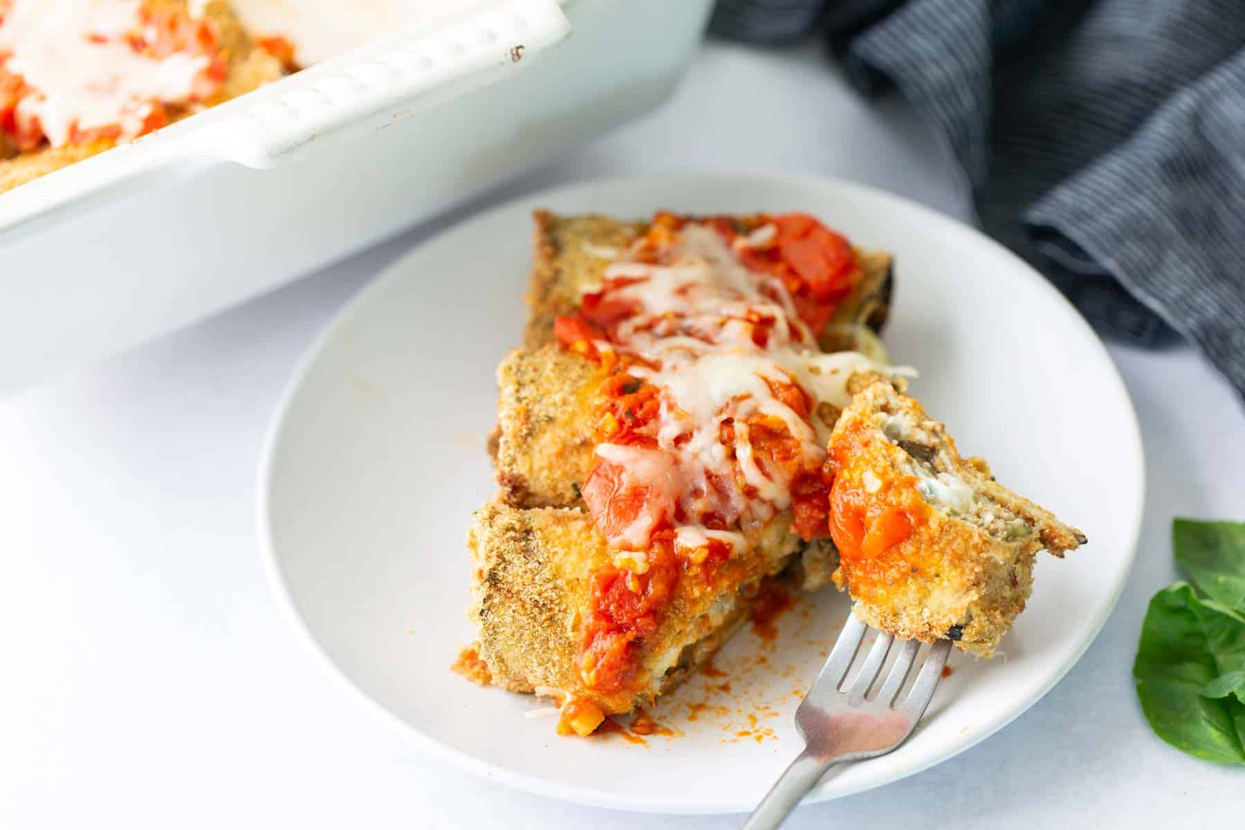 A white plate with breaded, baked eggplant slices topped with tomato sauce and melted cheese. A fork rests on the plate, and a baking dish and napkin are in the background.
