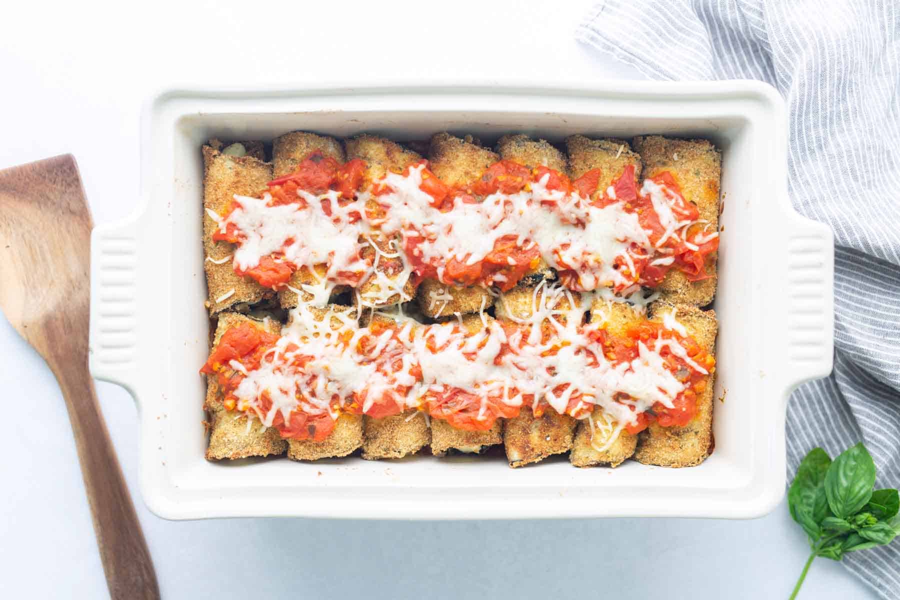 A white baking dish filled with breaded, rolled cannelloni topped with tomato sauce and melted cheese, next to a wooden spatula and a striped cloth.