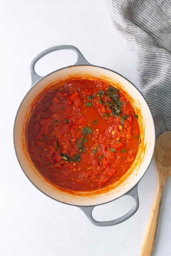 A pot of tomato sauce with chopped herbs sits on a white surface next to a wooden spoon and a striped cloth.